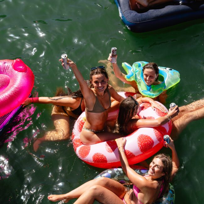 A group of five women in swimsuits enjoy a sunny day in the water, floating on colorful inflatable pool floats near a luxury yacht. They are raising their drinks and smiling at the camera. The water is a vibrant green, and the atmosphere is lively and cheerful, reminiscent of The Yacht Social Club Sydney boat hire experience.