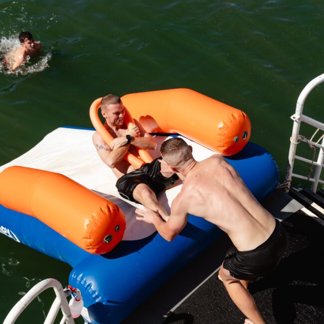 Two shirtless men are by the water. One is on an orange and blue inflatable float, while the other, standing on a dock, attempts to pull him off. Another person is swimming close by. The scene suggests playful interaction during a sunny day at a Luxury Yacht Rentals Sydney event by The Yacht Social Club.