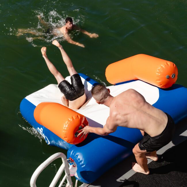 A shirtless man on a floating inflatable platform pushes a person headfirst into a lake while another swims nearby. Another person stands on the platform, watching the scene unfold at The Yacht Social Club Sydney Boat Hire.