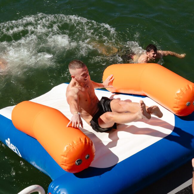 A person is about to slide off an inflatable water mat with orange handles, into a body of water. Another person is swimming nearby. The bright, sunny day enhances the vibrancy of the water and inflatables. The atmosphere appears fun and recreational.