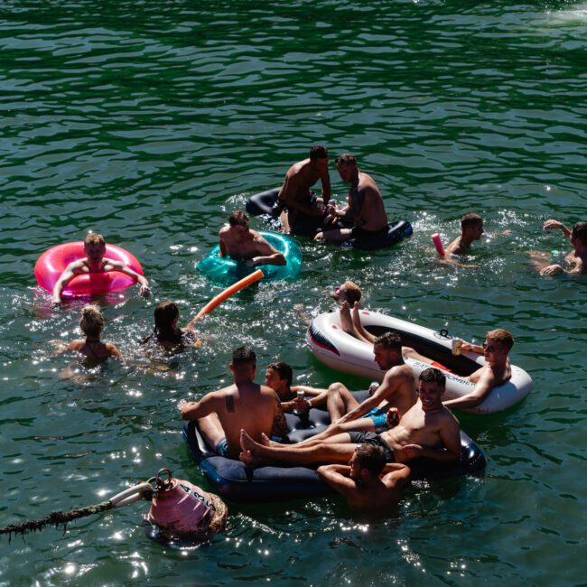 A group of people enjoying a day in the water, floating on various inflatable rafts and inner tubes. The water is green, and the weather appears sunny. People are socializing and relaxing, having a good time in their swimwear.