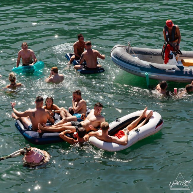 A group of people relaxes in inflatable rafts and floats on a sunlit body of water. A few individuals swim nearby, and others are in a small inflatable motorboat. The scene is lively and festive, suggesting a social gathering or party on the water.