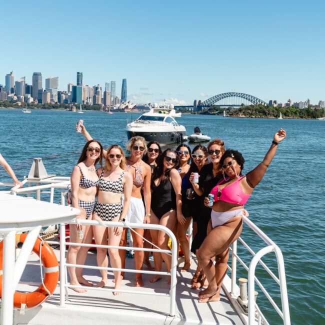 A group of nine women in swimwear pose and smile on a boat. The backdrop features a city skyline, including a bridge and several high-rise buildings, on a sunny day. The water is calm, and one woman waves her arm in the air enthusiastically.