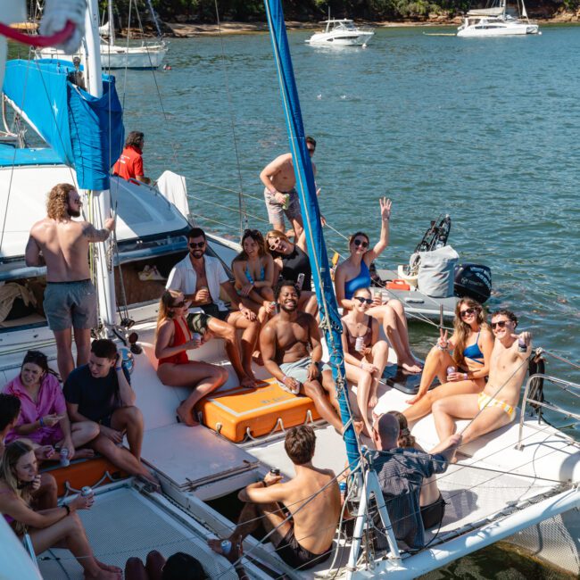 A group of people are relaxing and socializing on a catamaran in a scenic bay. Some are sitting and lying on the deck while others are standing. The background features calm waters, lush green trees, and a few other boats. It is a sunny day.