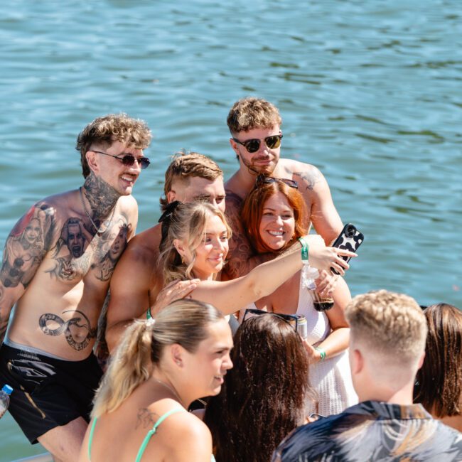 A group of people, some with tattoos and sunglasses, are gathered closely together on a boat, smiling and posing for a selfie. They are dressed in summer attire with a scenic background of blue water. The sunny weather suggests a fun and lively outdoor gathering.