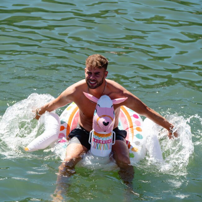 A smiling shirtless man with short hair and a beard is sitting on an inflatable llama float in the water. He is wearing dark swim trunks and splashing water with his hands. The water around him appears calm and greenish.