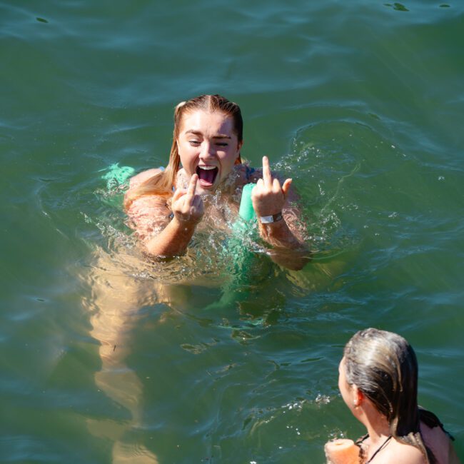 Two people are in the water, with the individual in the foreground facing away from the camera. The individual in the background with long hair and a green swimsuit is mid-swim, making a gesture with both hands and smiling broadly.