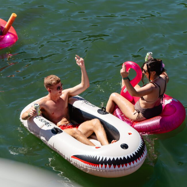 Two people are floating on inflatable pool toys in a body of water. The person on the left is in a white inflatable with a shark design, holding a drink, and raising an arm. The person on the right is on a pink flamingo inflatable, also holding a drink.