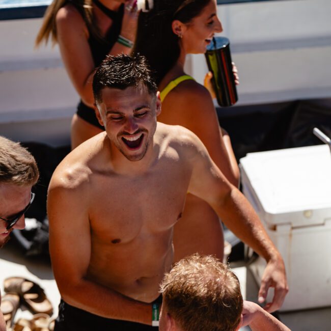 A group of people are socializing on a boat. A shirtless man in the center is laughing, surrounded by others who seem to be talking and having a good time. The atmosphere appears casual and festive. There are drinks and coolers visible on the boat.