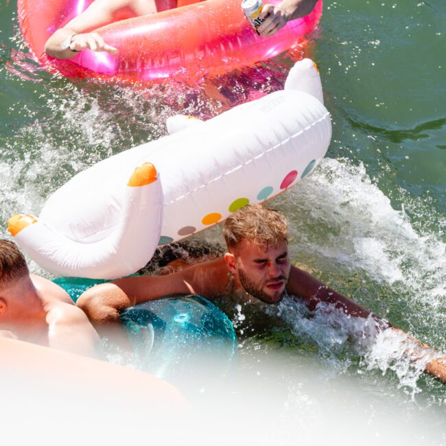 Two men are in a body of water with inflatable pool floats. One man with a beard is partially submerged, holding onto a large inflatable that resembles a white dog. The other man holds a red float nearby. Water splashes around both as they enjoy their time.