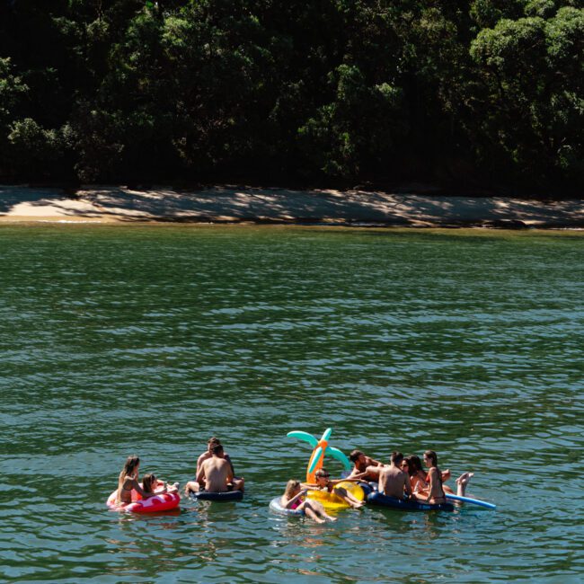 A group of people relax on inflatable floaties in a calm lake surrounded by dense greenery. The sun shines brightly, creating a peaceful, outdoor recreational scene.