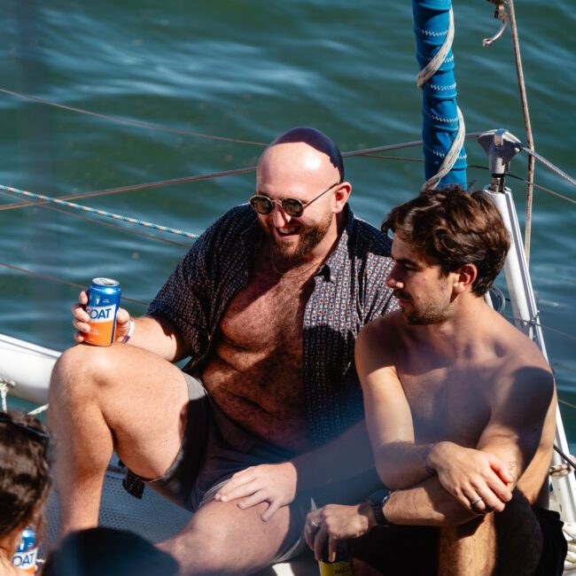 A group of people enjoy a sunny day on a sailing boat. A bald man wearing sunglasses and holding a can sits next to another man with wavy hair. Several others are seated around, soaking up the sun and socializing. The sea is visible in the background.