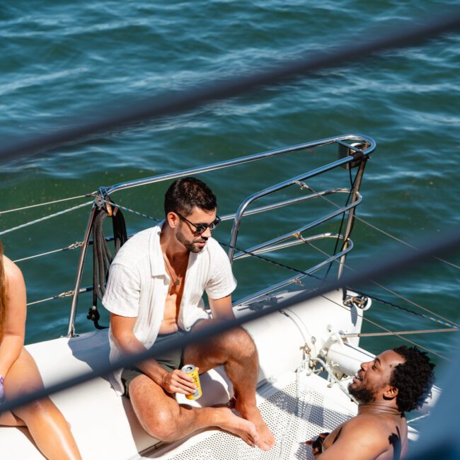 A group of people relax on a boat under the sun, with two men seated at the front. One man holds a drink while chatting, wearing sunglasses and casual attire. The water behind the boat is calm and clear, and a logo in the bottom right corner reads "Yacht Social.