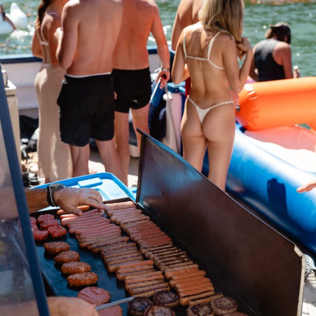 People in swimsuits gather near a barbecue grill loaded with burgers and hot dogs on a sunny day by the water. The scene includes inflatable water toys in the background, suggesting a summer party or gathering by the lake or pool.