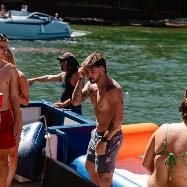 A group of people enjoys a sunny day by the water. Several shirtless men and women in swimsuits are socializing on and around an inflatable raft. In the background, a boat is anchored, and a man in a hat stands waist-deep in the water. The setting is lively and relaxed.