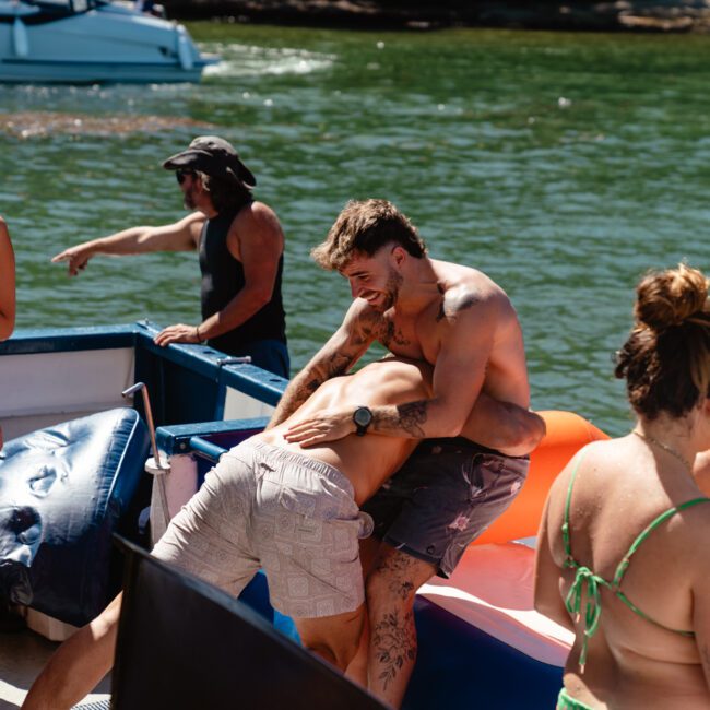 A group of people on a boat. Two shirtless men are playfully wrestling at the edge. A shirtless man in red shorts stands nearby holding a red cup. Other people, including a woman in a green bikini, are standing around or sitting on orange inflatables. The background shows water and trees.