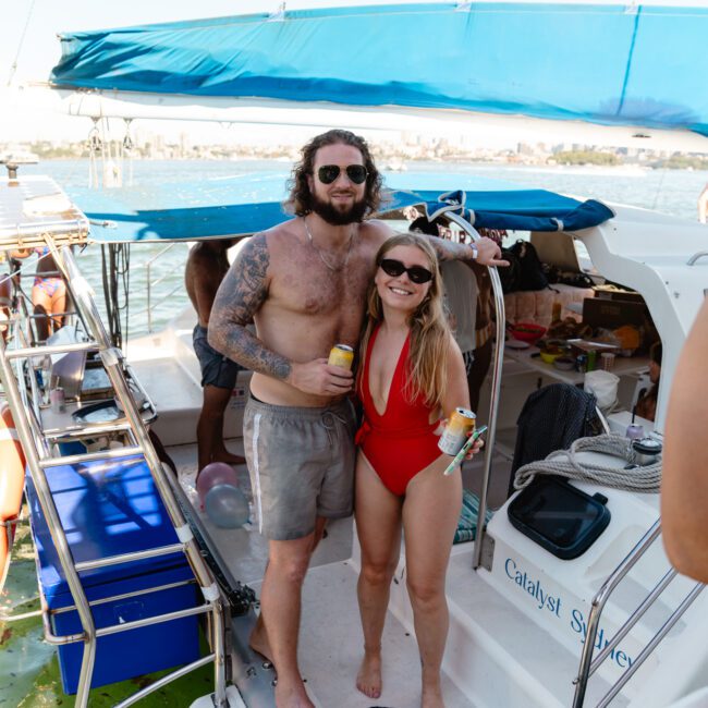 A shirtless man with sunglasses and tattoos poses alongside a woman in a red swimsuit and sunglasses on a boat. Both are holding drinks and smiling. The boat, named Catalyst Survey, is docked near a body of water with a background of distant buildings.