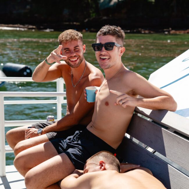 Three shirtless men are relaxing on a boat. Two are sitting on a bench, smiling at the camera, one wearing sunglasses and holding a cup, while the third is lying down on the bench, face down. Behind them is a body of water and a partially visible shoreline with trees.