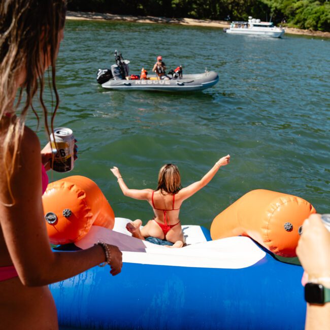 A woman in a red bikini sits on an inflatable slide, about to enter the water. She has her arms raised and is facing away from the camera. Another woman holding a drink stands nearby. A small boat with people is on the water in the background. Lush greenery surrounds the scene.