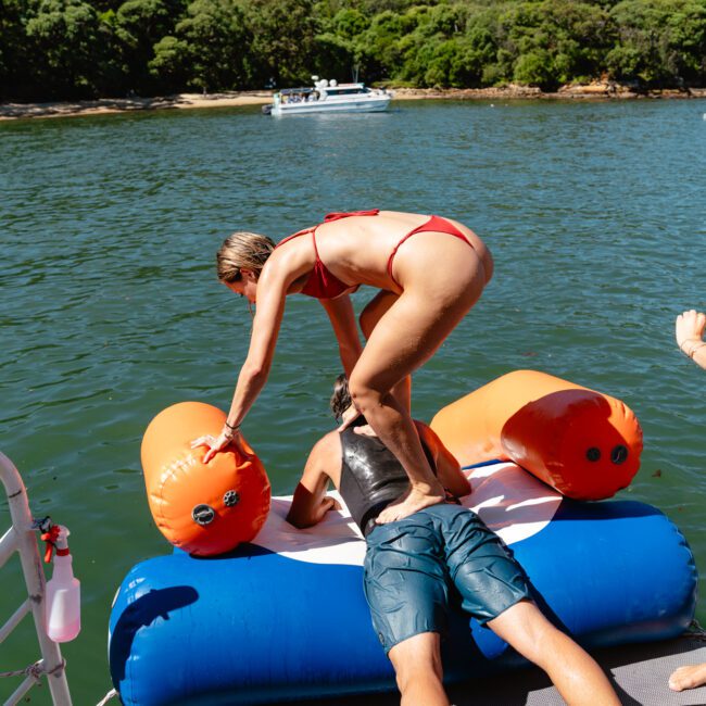 A woman in a red bikini is gently stepping over a person lying facedown on an inflatable water float. The inflatable is orange and blue, situated near a dock by a lake. In the background, there is a forested shoreline and a boat on the water.