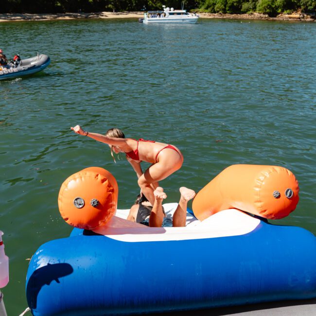 A person in a red swimsuit playfully pushes another person, who is lying on an inflated water float, into a lake. There are trees and other boats in the background under clear skies, suggesting a sunny day. The scene captures a fun moment by the water.