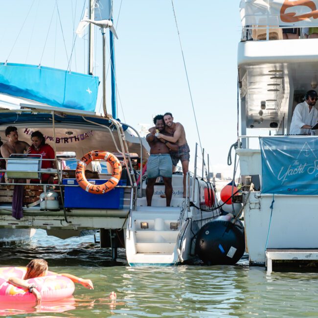 People are enjoying a sunny day on two docked boats. One person is floating in an inflatable pink tube in the water. A couple hugs on the boat walkway while others are relaxing and socializing on the boats. A banner reads "Yacht Social.