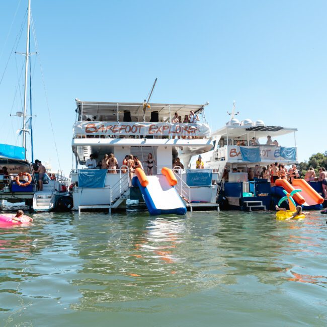 A group of people enjoying a sunny day on two large boats docked side by side, equipped with water slides leading into a lake. Some people are swimming or floating on inflatable rings in the water, while others are relaxing or socializing on the boats. Trees and part of a city skyline are visible in the background.