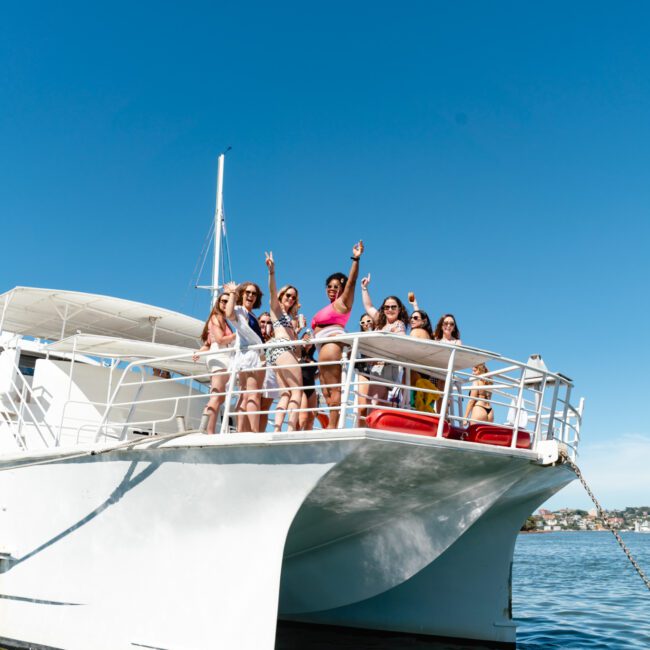 A group of people stands on the deck of a catamaran under a clear blue sky. They are smiling, with some posing and others cheering with their hands raised. The water and distant shoreline are visible in the background. The boat has "Yacht Social Club" branding.