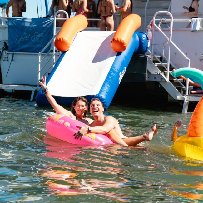 Two people are happily floating on a pink inflatable tube in the water, with others in the background near a boat with a slide. Another inflatable, shaped like a palm tree, is nearby. The atmosphere is lively and joyous.