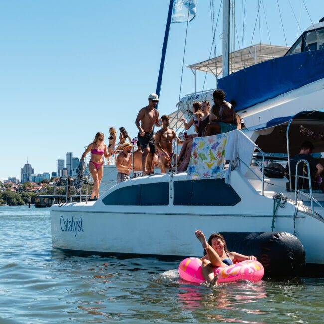 A group of people enjoy a sunny day on a catamaran named "Catalyst." Some participants are standing on the deck, while a woman floats in the water on an inflatable ring. The city skyline is visible in the background, and the water is calm.