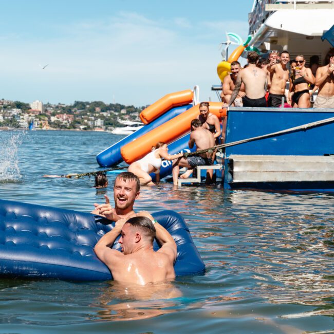 A group of people enjoy a sunny day on the water. Two men are smiling and playing with a blue inflatable mattress in the water near a boat, while others on the deck watch and have fun. Several inflatables are in the water, and the background shows trees and buildings.