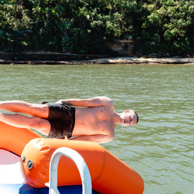 A shirtless person in black shorts is lying sideways on an orange inflatable water toy. The toy is positioned near the edge of a body of water, with a forested area visible in the background. There is a metal ladder in the foreground.