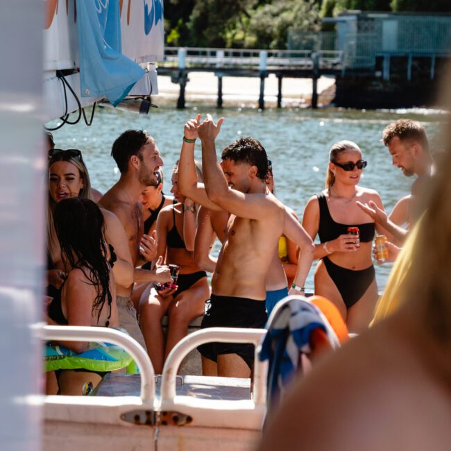 A group of people enjoying a sunny day on a boat, some standing and others sitting. Two shirtless men are dancing, while others are chatting and holding drinks. The boat is near a dock, and trees and water are visible in the background.