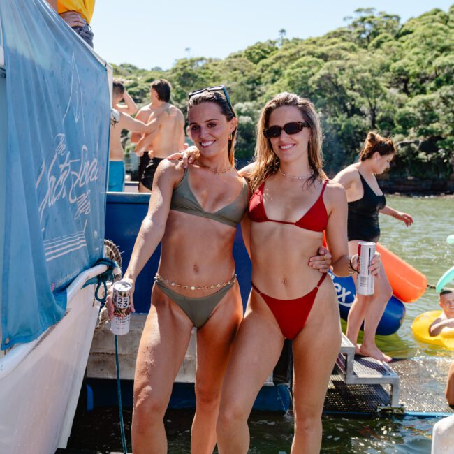 Two women in bikinis—one in olive green and the other in red—smile while holding drinks and posing on a boat. The boat is docked near a lush, green shoreline with other people enjoying the sunny day in the background.