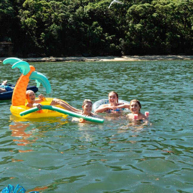 Four people enjoy themselves in a lake surrounded by trees, with two of them on an inflatable palm tree floatie, another on a blue pool float, and the fourth person holding onto the floatie. The water is green and a sunny, clear day is evident.