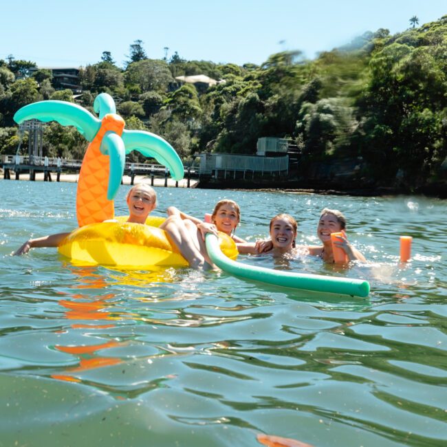 Four children smiling and playing in the water with a floating inflatable palm tree and pool noodles on a sunny day. Trees and a dock are visible in the background.