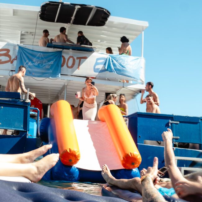 People are enjoying a party on and around a boat. Some are on an inflatable slide attached to the boat, while others relax in the water. The boat's upper deck has people socializing and lounging, with banners hanging on the sides. The atmosphere is lively and fun.