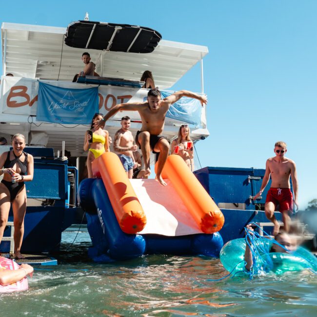 A group of people enjoys a sunny day on a boat with a slide. One person is mid-air after sliding into the water, while others watch and swim nearby. The boat features banners reading "Yacht Social." The water is clear, and everyone seems to be having fun.