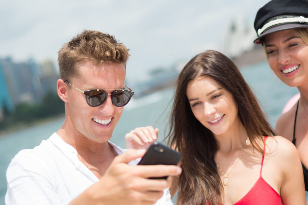 Three people standing outdoors near the water, smiling and looking at a smartphone. The man on the left wears sunglasses and a white shirt, while the two women on the right are in summer outfits, one wearing a captain's hat. Cityscape and water are visible in the background, possibly hinting at The Yacht Social Club Sydney Boat Hire experience.