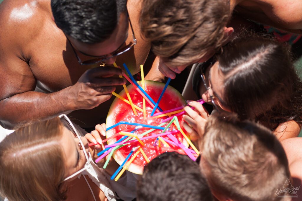 A group of people gathered closely together are sipping from colorful straws placed into a halved watermelon. The watermelon is filled with a liquid, and the straws are inserted around the edge and middle. This casual and fun scene perfectly captures the vibe of Boat Parties Sydney with The Yacht Social Club.