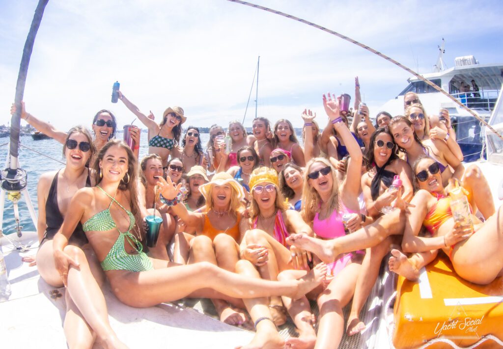 A large group of people smiling and posing for a photo on a boat during The Yacht Social Club event. Many are wearing swimsuits and sunglasses, holding drinks, and making celebratory gestures. The sun is shining overhead, with the water and another boat visible in the background.