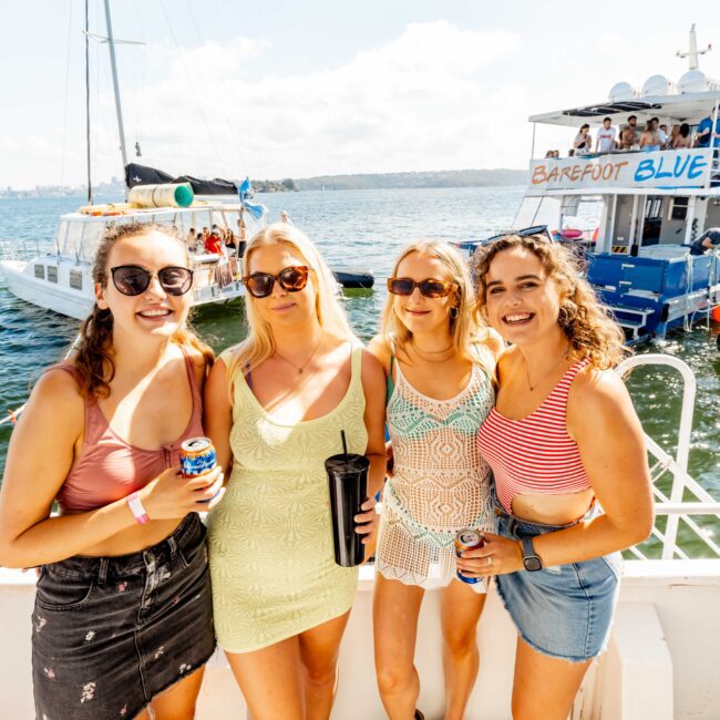 Four women, dressed in summer attire, stand on a boat with the ocean and another party boat labeled "Barefoot Blue" visible in the background. They are smiling, holding drinks, and wearing sunglasses on a sunny day. Enjoying Boat Parties Sydney The Yacht Social Club, several other boats are also visible on the water.