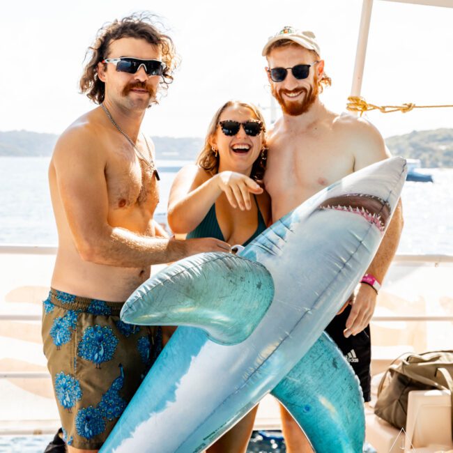 Three people in swimwear are posing on a boat, holding a large inflatable shark. Two men in sunglasses are on either side of a woman wearing sunglasses and smiling broadly. They are enjoying a sunny day out on the water with coastal scenery in the background, courtesy of The Yacht Social Club.