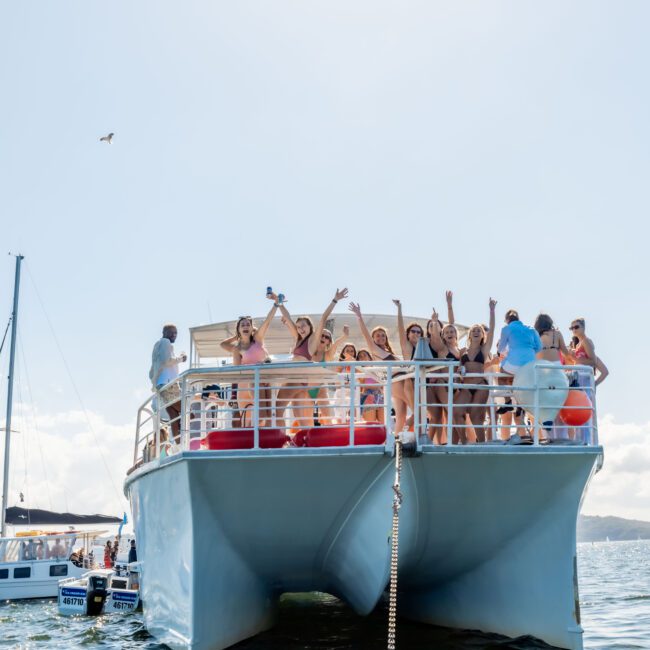 A large group of people are partying on the upper deck of a catamaran anchored in a calm body of water. Other boats and a mountainscape can be seen in the background under a sunny sky. The Yacht Social Club Event Boat Charters provide an unforgettable experience for boat parties in Sydney.