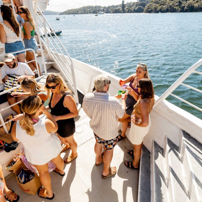 A group of people enjoying a sunny day on a boat. Some are sitting while others stand, conversing and holding drinks. The yacht is docked near a scenic shore with lush greenery and calm waters in the background, perfect for events like those by The Yacht Social Club Sydney Boat Hire.