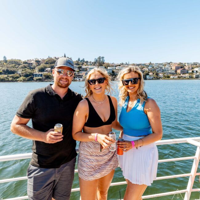 Three people smiling and posing on a boat with clear blue water and a hilly, residential shoreline in the background. They are wearing sunglasses and holding drinks, enjoying a sunny day. Two women are in summer attire, and the man is in a black shirt and shorts. Experience it with The Yacht Social Club Sydney Boat Hire!