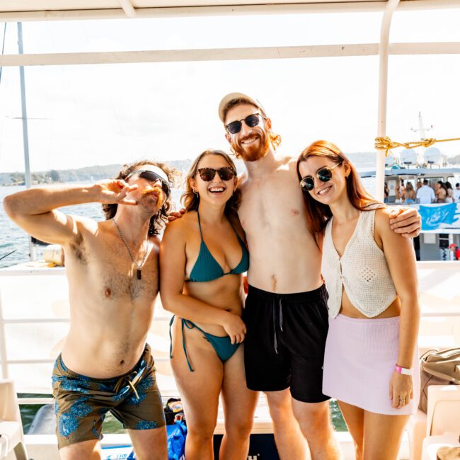 Four people posing and smiling on a boat deck on a sunny day. One man is making a peace sign, one woman is wearing a blue bikini, another man is shirtless and wearing sunglasses, and a second woman is in a white crop top and pink skirt. A body of water and another boat are in the background, capturing the vibe of the Yacht Social Club Event Boat Charters.