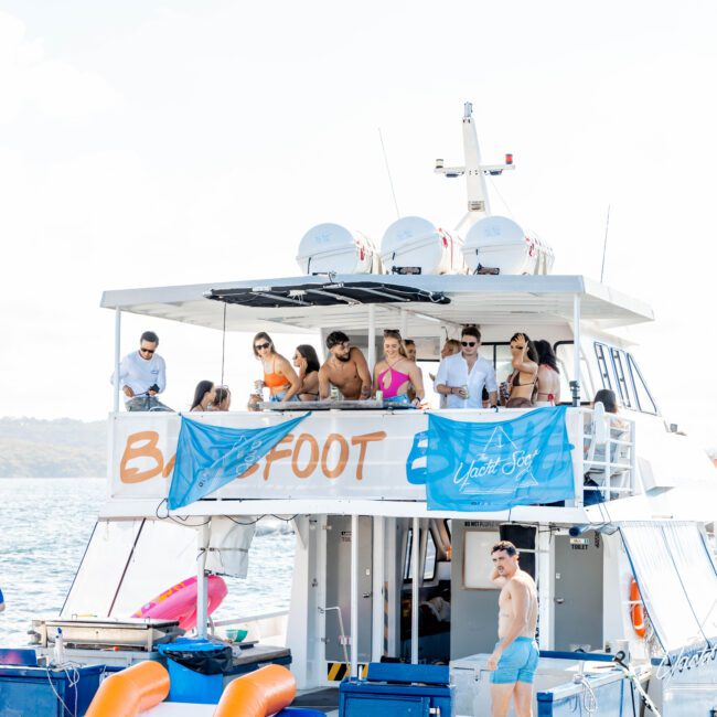 A group of people enjoying a sunny day on a two-story boat named "Barefoot" in Sydney Harbour. Some are on the upper deck, leaning over the railing, while others are on the lower deck and in the water nearby. The boat has blue banners and several orange floats drifting around, making it perfect for parties with The Yacht Social Club.