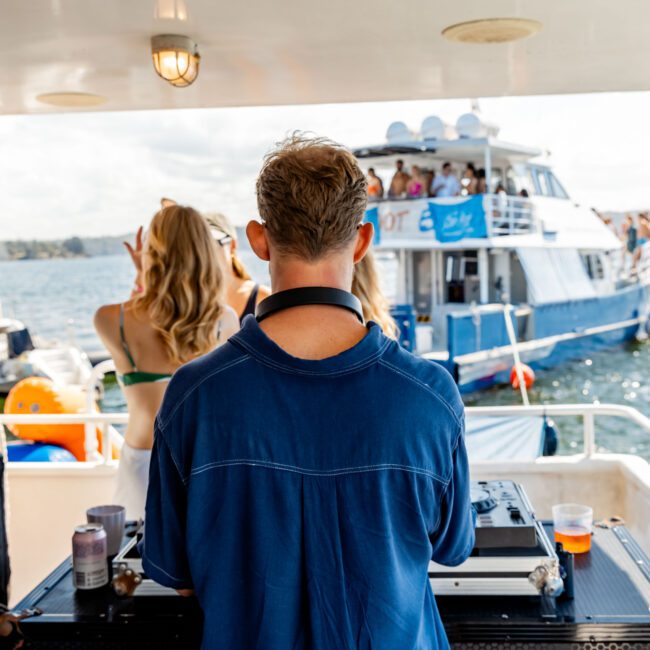 A man in a blue shirt is DJing on a boat, facing a turntable setup. A blonde woman in a swimsuit dances nearby. Another boat with people onboard is visible in the background on a sunny day over the water, part of The Yacht Social Club Sydney Boat Hire event.