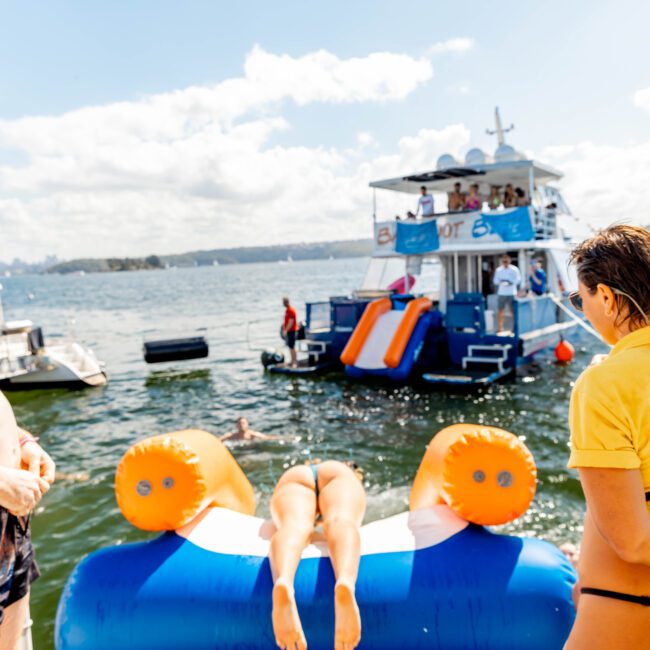 A group of people in swimsuits enjoys a sunny day on the water. One person is on an inflatable tube, ready to be launched, while others watch. In the background, there's a yacht from The Yacht Social Club with slides and more people relaxing. The scene is lively and festive, perfect for Boat Parties Sydney.